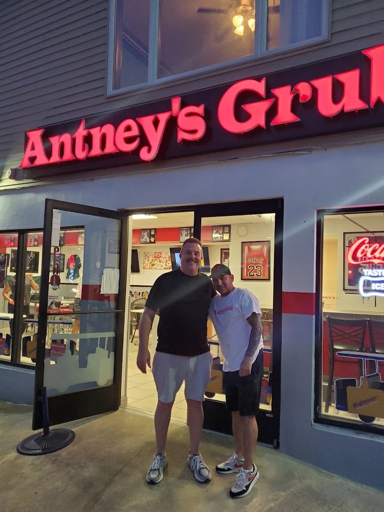 Two people standing outside Antney's Grub at night in front of the illuminated sign